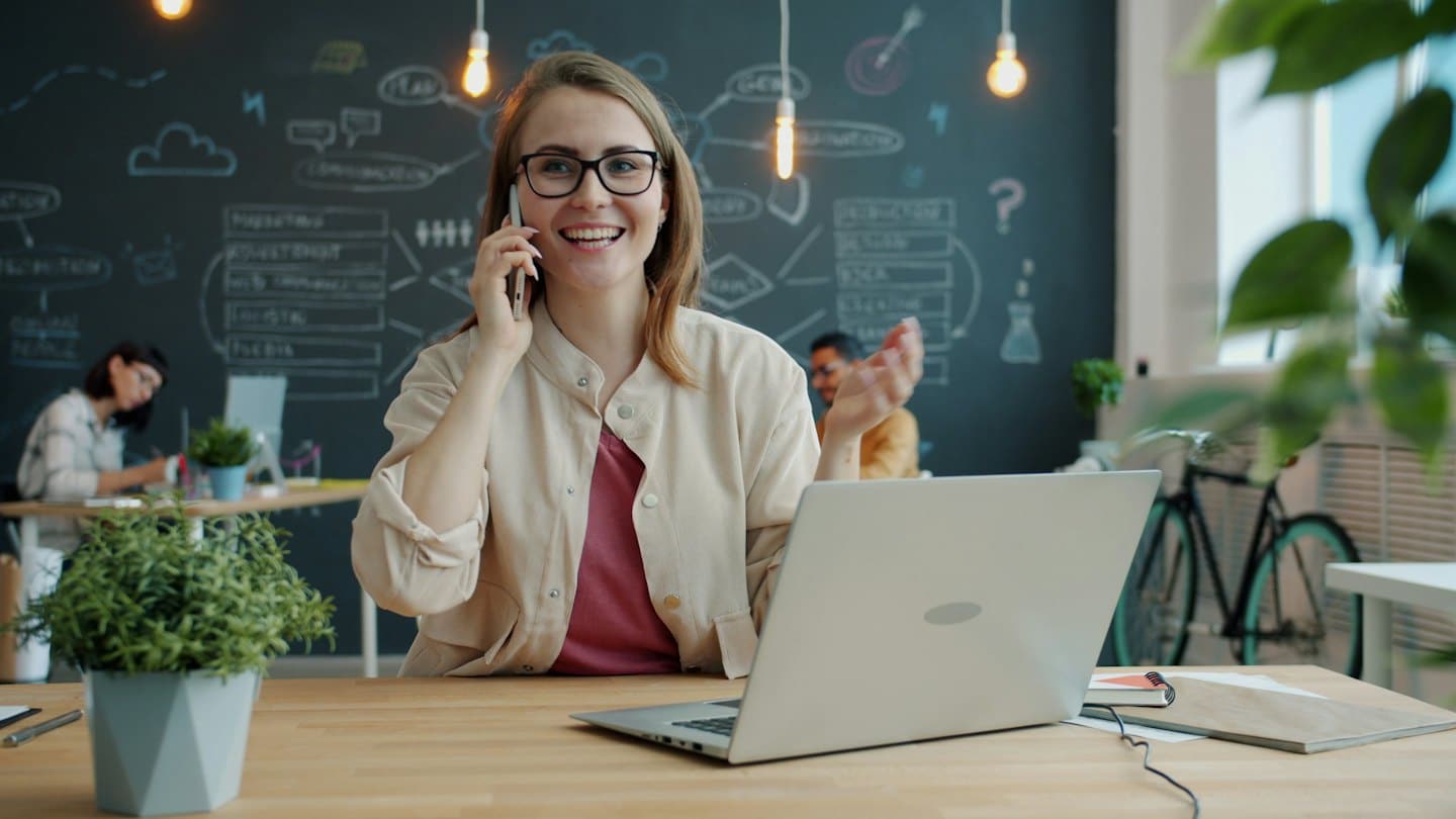 Mujer con gafas habla por teléfono en una oficina moderna.